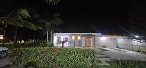 A modern house with a well-lit entrance, a stone wall, and a garden with red flowers. - Villa La Escapada - Private Caribbean Oasis Near Juan Dolio Beach (San Pedro de Macorís)