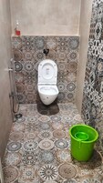A bathroom with a toilet, patterned tiles, a green bucket, and a soap dispenser.