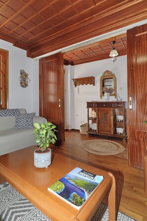 A living room with a wooden ceiling, a sofa, a coffee table with a magazine, a potted plant, and a wooden cabinet with a mirror. - Magdalini's Holiday Home in Skopelos (Skopelos)
