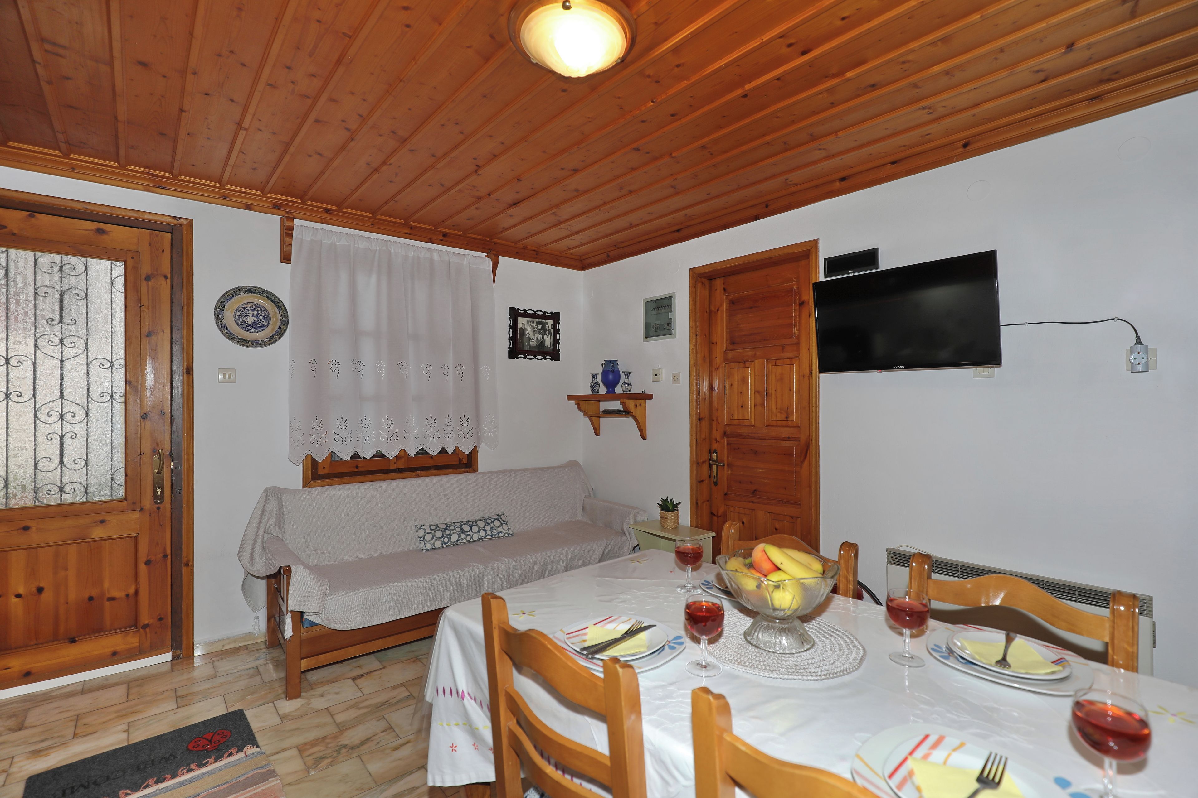 A dining area with a wooden table set for two, a television mounted on the wall, and a wooden door.