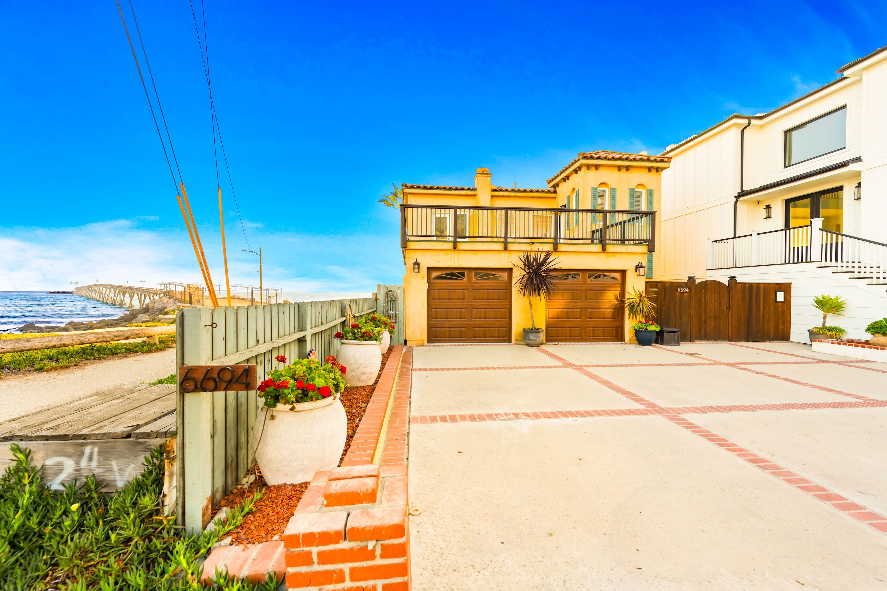 A coastal residential area with a two-story house featuring a balcony, a wooden fence, and a paved driveway.