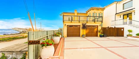 Un área residencial costera con una casa de dos pisos que tiene un balcón, una valla de madera y un camino de entrada pavimentado.