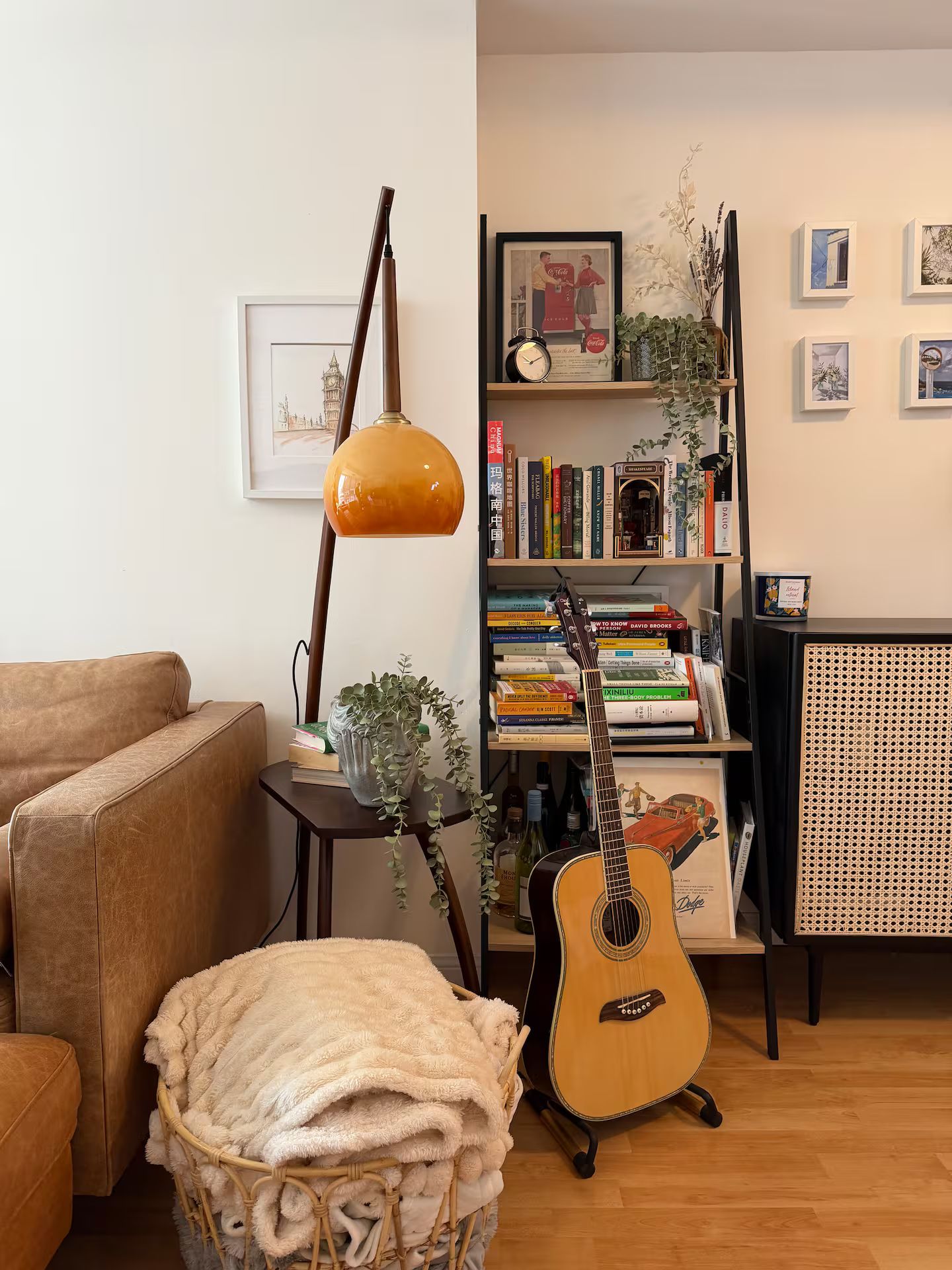 A cozy living room with a brown leather sofa, a white fluffy throw, a guitar, a bookshelf filled with books, and a decorative lamp.