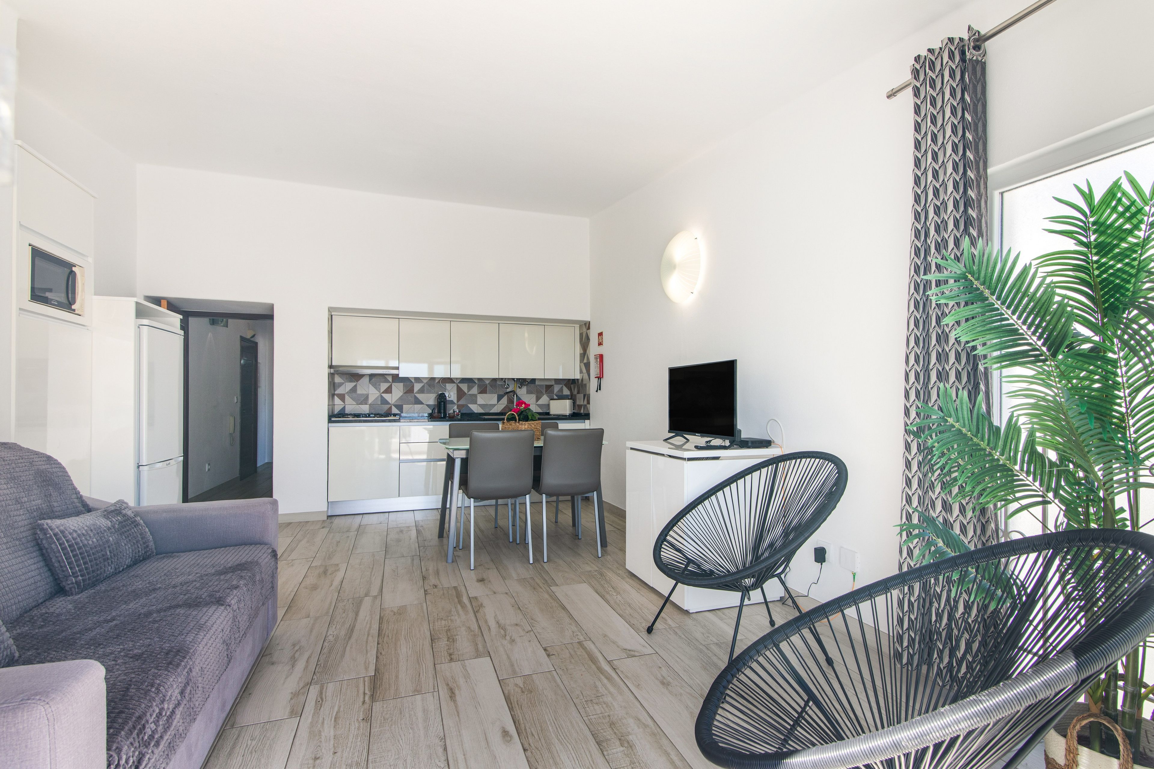 Una sala de estar moderna con un sofá gris, un comedor con mesa y sillas, una cocina con armarios blancos y un televisor sobre un mueble blanco.