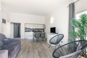 Ein modernes Wohnzimmer mit grauem Sofa, einem Essbereich mit Tisch und Stühlen, einer Küche mit weißen Schränken und einem Fernsehgerät auf einem weißen Lowboard.