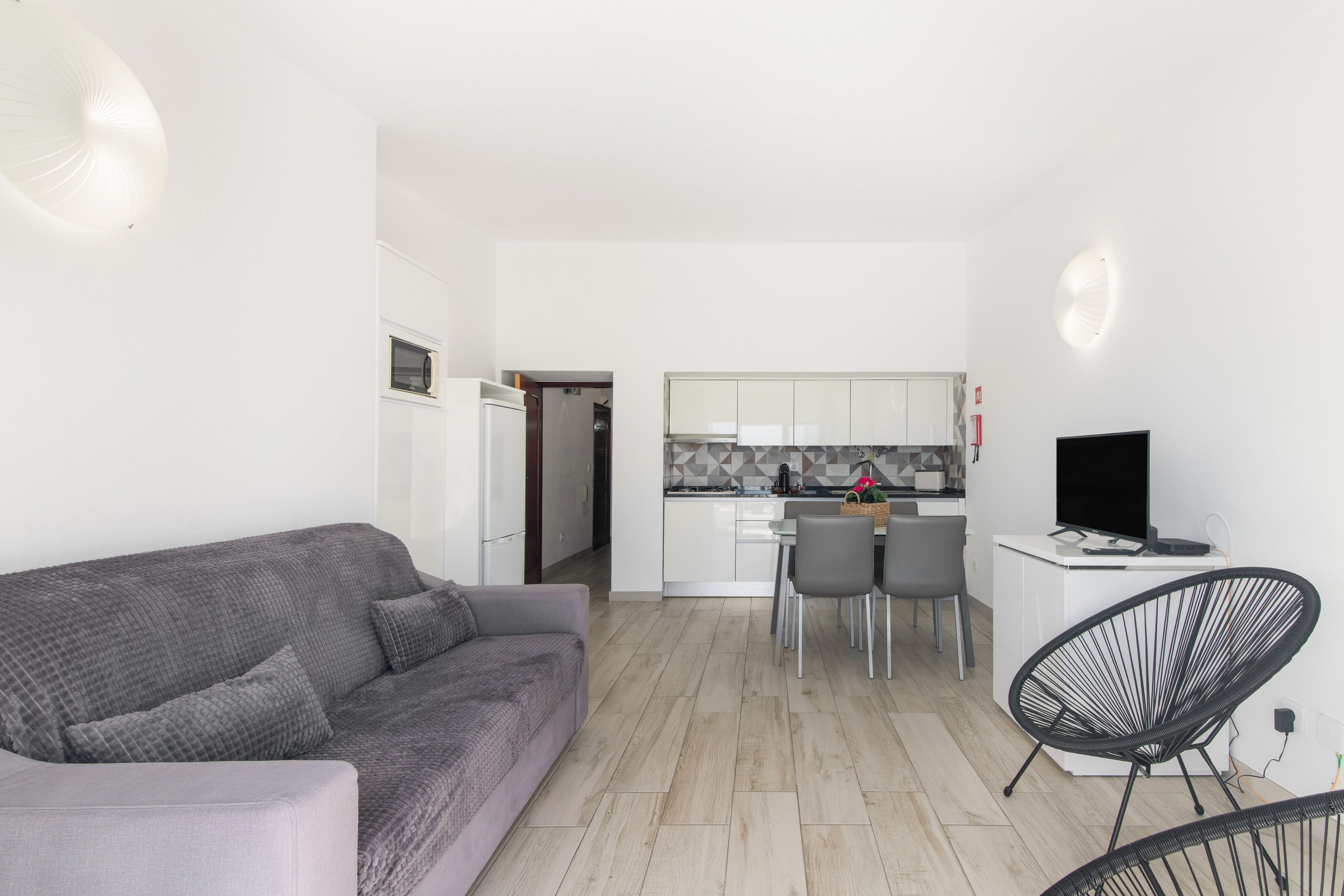 Una sala moderna con un sofá gris, un comedor con mesa y sillas, y una cocina con armarios blancos y un televisor negro.
