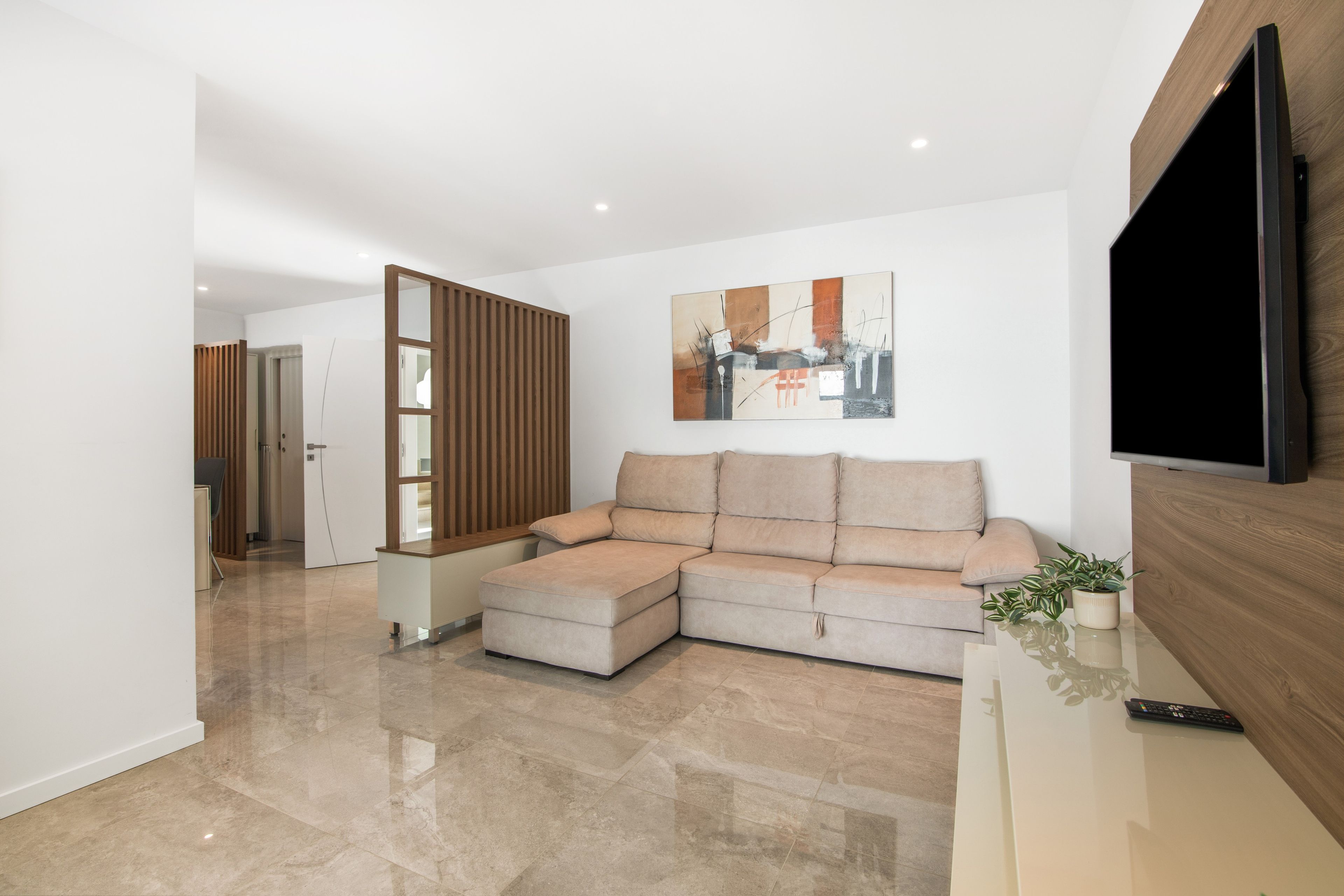 Een moderne woonkamer met een hoekbank, een flatscreen televisie aan de muur en een houten scheidingswand.