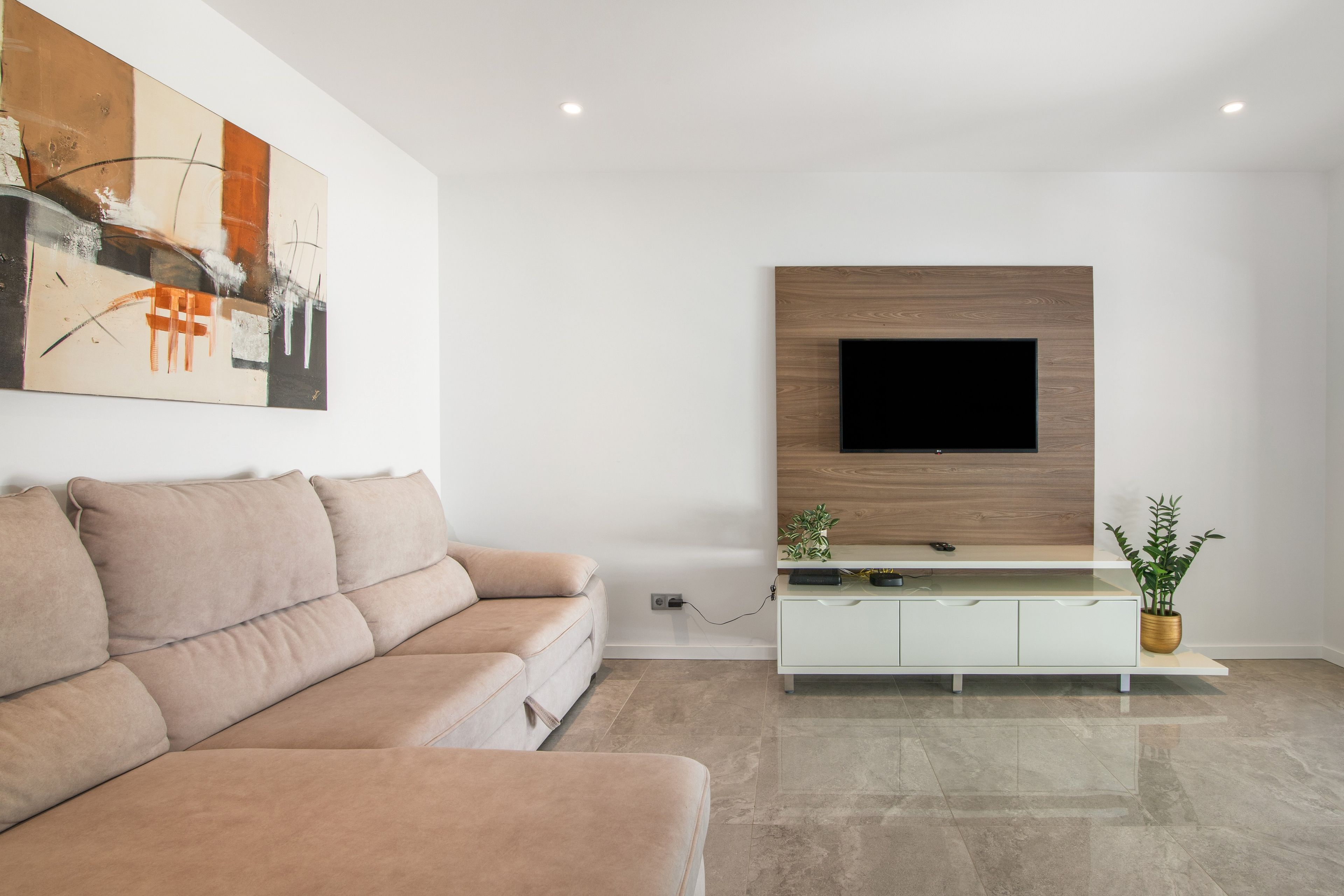 Un salon moderne avec un canapé beige, un téléviseur à écran plat fixé sur un panneau en bois et un meuble blanc orné d’une plante.