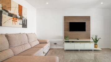 Un salon moderne avec un canapé beige, un téléviseur à écran plat fixé sur un panneau en bois et un meuble blanc orné d’une plante.