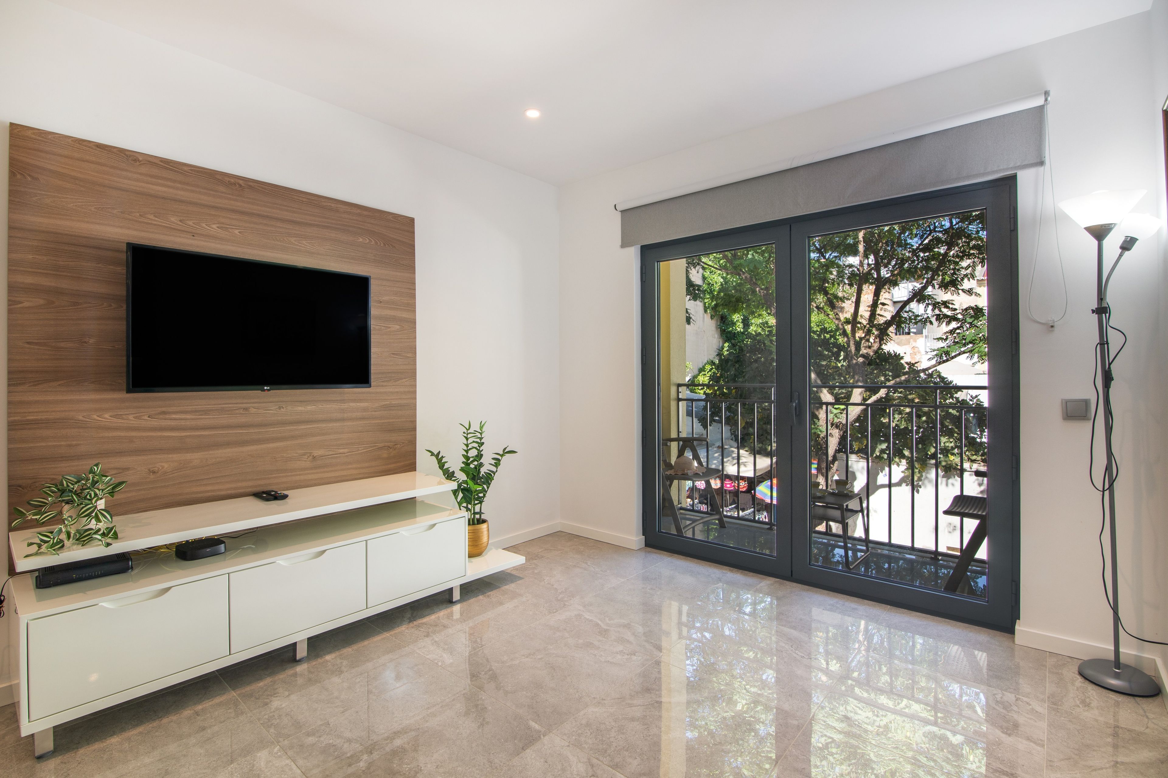 Een moderne woonkamer met een flatscreen televisie aan een houten paneel, een wit tv-meubel, een potplant en een schuifdeur naar een buitenruimte.