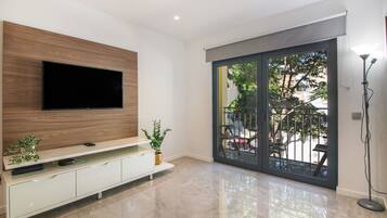 Een moderne woonkamer met een flatscreen televisie aan een houten paneel, een wit tv-meubel, een potplant en een schuifdeur naar een buitenruimte.