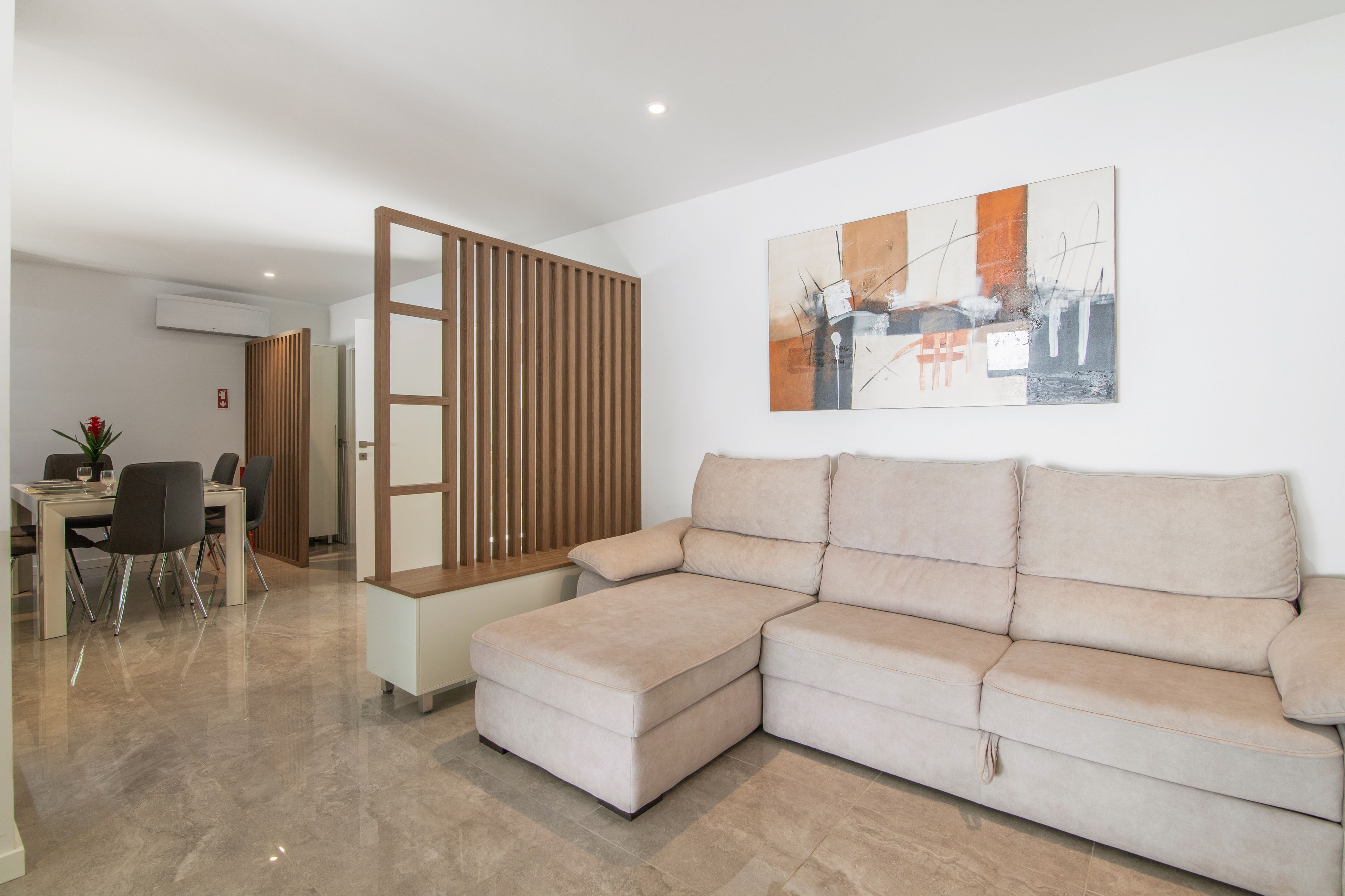 A modern living room with a sectional sofa, a dining area with chairs, and a wall-mounted air conditioner.