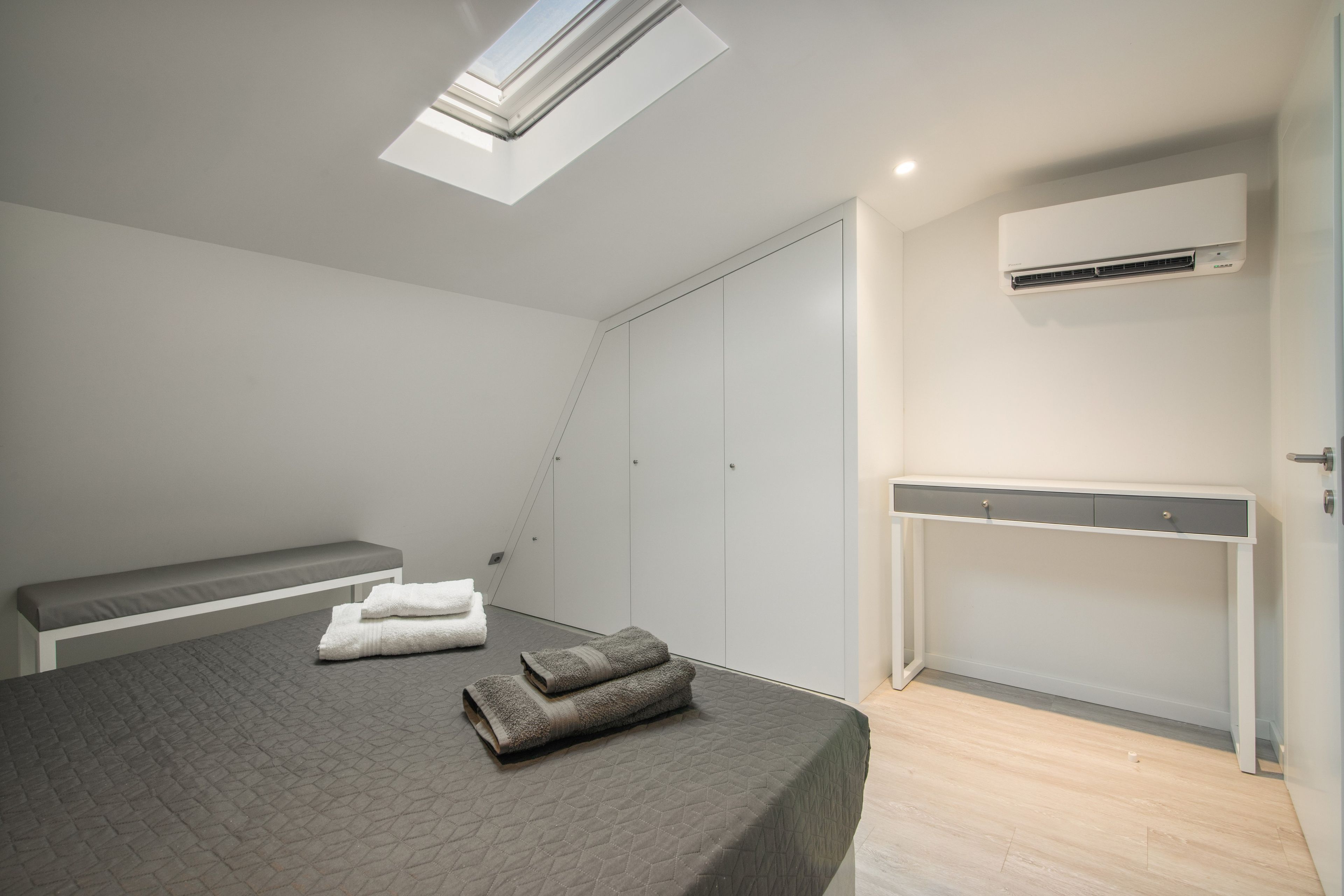 Une chambre à coucher moderne avec un puits de lumière, un lit recouvert d’une courtepointe matelassée, un banc, une armoire et un appareil de climatisation.