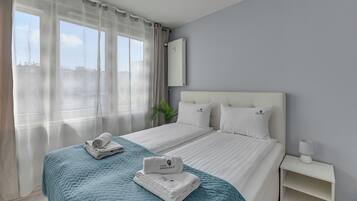 Quarto de hotel com cama, mesa de cabeceira, abajur, janela com cortinas e vista para prédios.