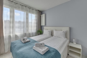Habitación de hotel con cama, mesita de noche, lámpara, ventana con cortinas y vista a edificios.