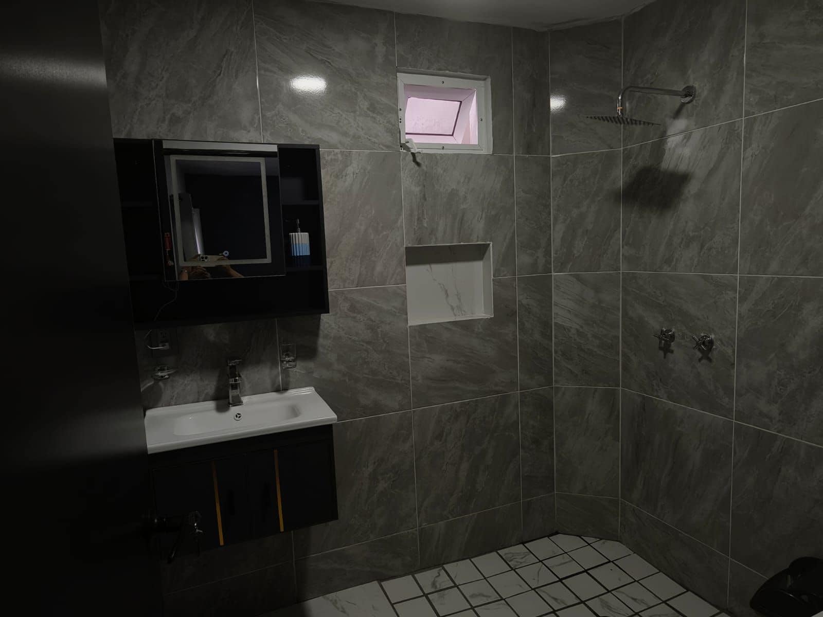A modern bathroom with a built-in sink, a mirror, a shower, and a small window.