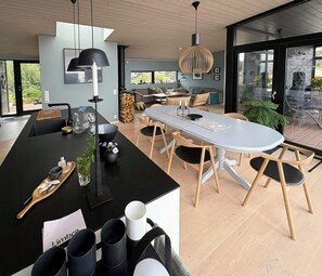 A modern kitchen with a central island, dining area with a round table, and a large window providing a view of the outdoors. - Fjord Idyll – Peace, Nature & Views (Vestland)
