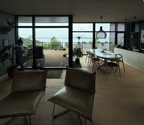 A modern interior with a dining area, two leather chairs, a table, and a view of the sea through large windows.
