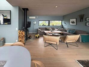 A modern living room with a sofa, coffee table, and chairs. There is a fireplace with a TV, and a window with a view of greenery.