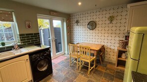 A kitchen with a washing machine, a wooden table with chairs, a clock on the wall, and a patterned wallpaper. - Cosy cottage in Wakefield. Perfect for long stays (Wrenthorpe)