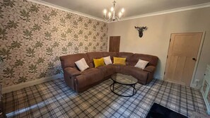 A living room with a brown leather sofa, a glass coffee table, plaid carpet, floral wallpaper, a chandelier, and a mounted deer head. - Cosy cottage in Wakefield. Perfect for long stays (Wrenthorpe)
