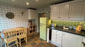 Una cocina con papel tapiz estampado, un reloj, un refrigerador verde, una mesa de madera con una silla, y un letrero que indica un pub.