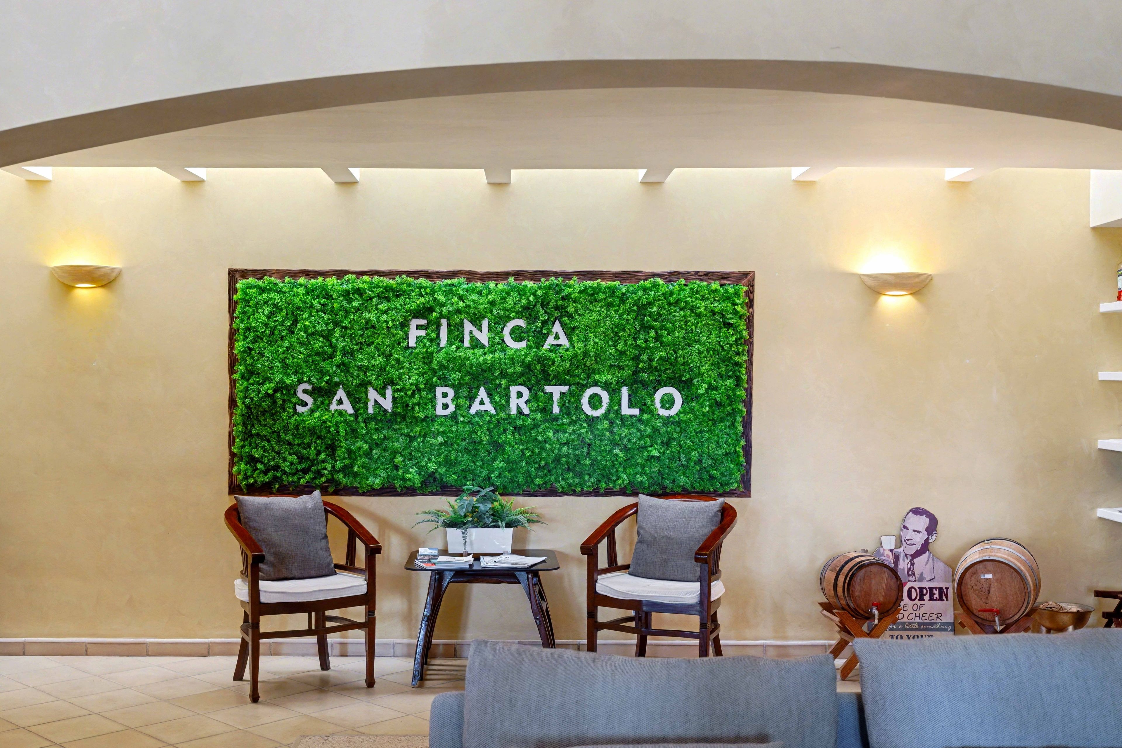 A reception area with a green wall displaying 'FINCA SAN BARTOLO' and two wooden chairs with cushions.