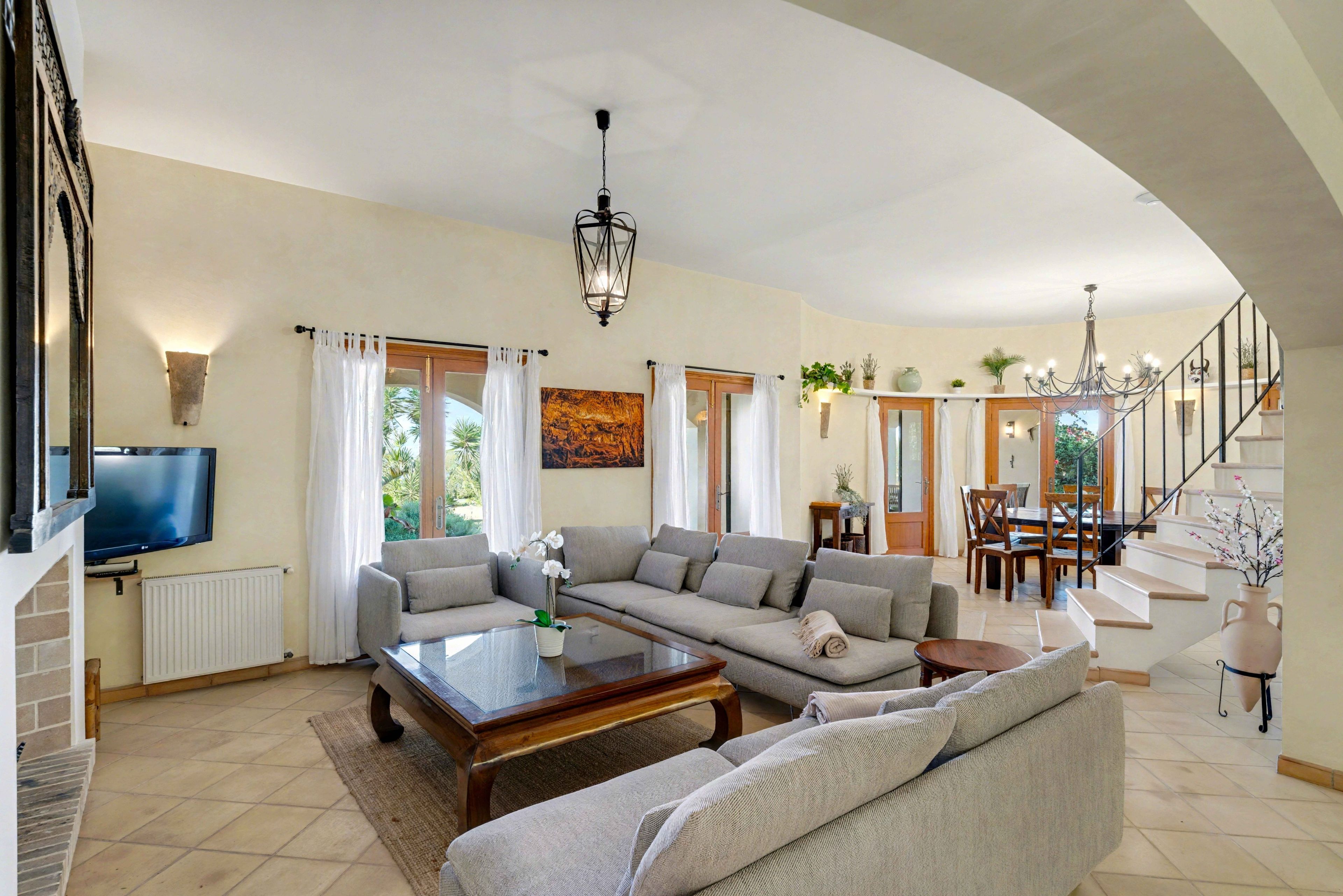 A spacious living room with a large sofa, a coffee table, and a TV. There is a staircase leading to an upper level.