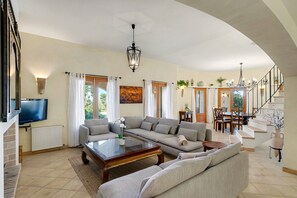 A spacious living room with a large sofa, a coffee table, and a TV. There is a staircase leading to an upper level.
