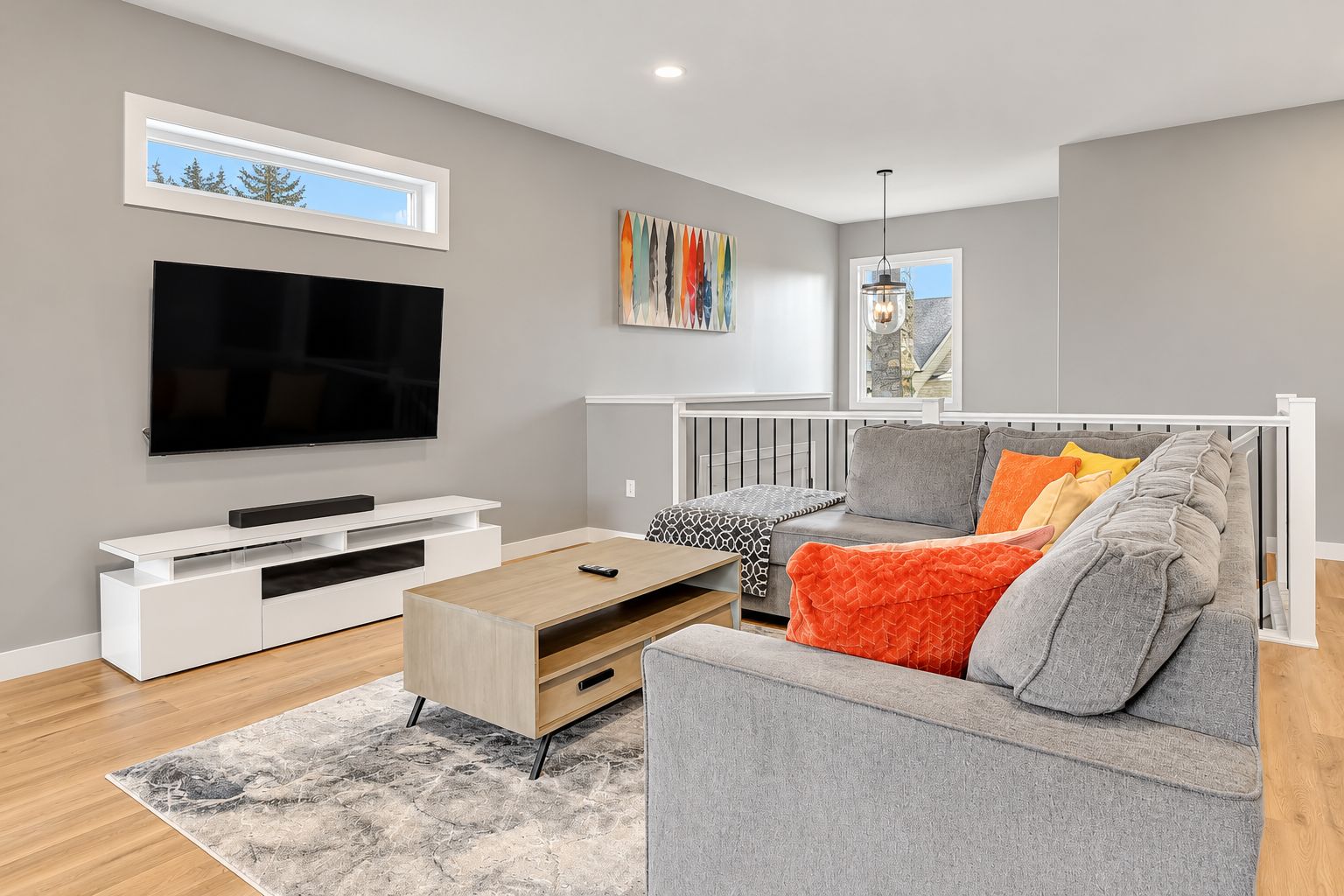 Un salon moderne avec un téléviseur à écran plat, un meuble de rangement blanc, un canapé gris agrémenté de coussins orange et gris, une table basse en bois et un tapis gris.
