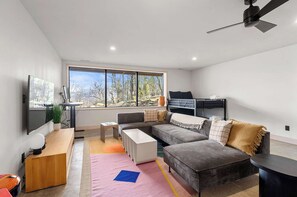 Ein modernes Wohnzimmer mit einer Couch, Couchtisch und Fernseher. Ein großes Fenster bietet Blick auf Bäume und einen Balkon.