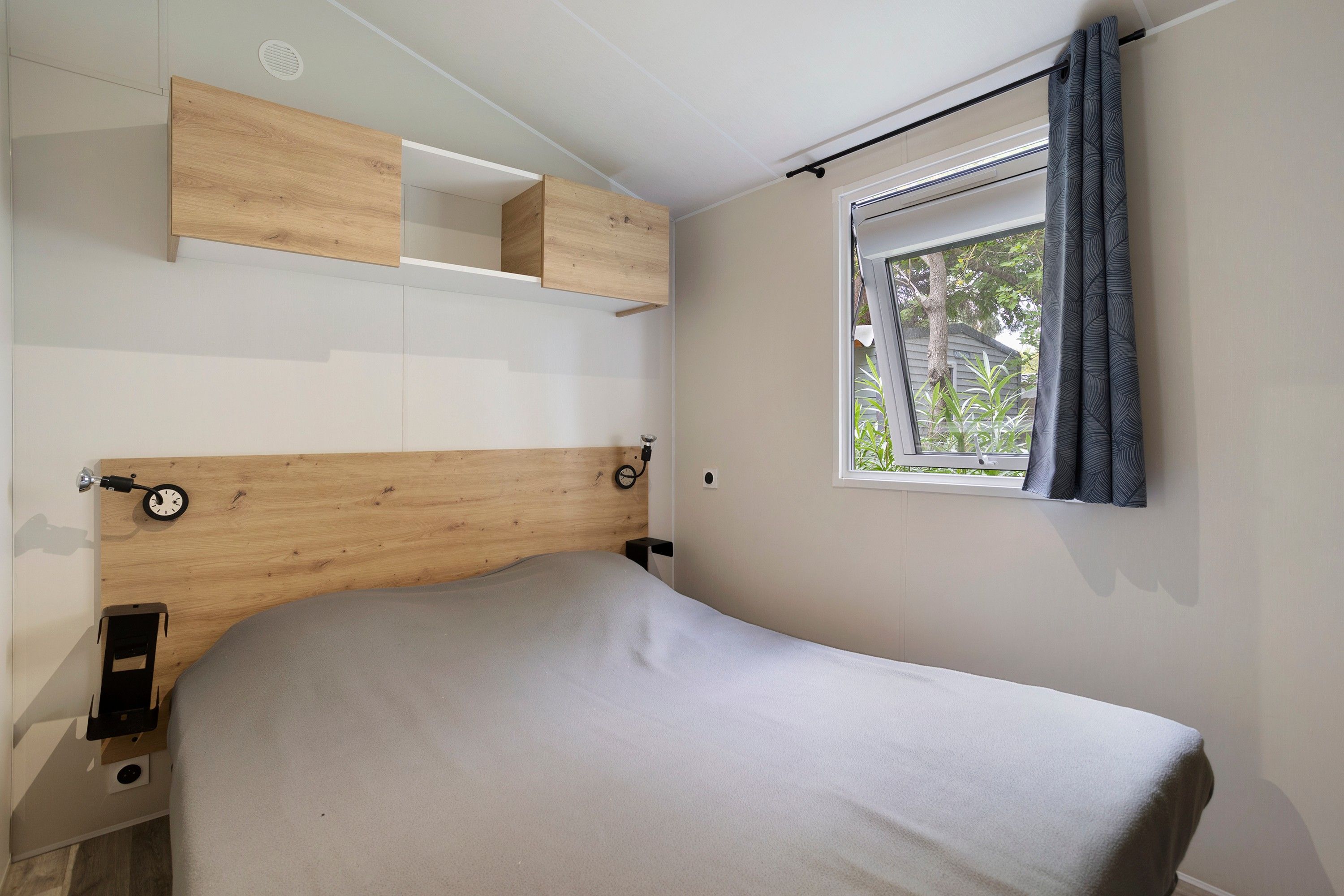 A bedroom with a wooden headboard, a bed with a grey cover, a window with curtains, and built-in shelving above the bed.