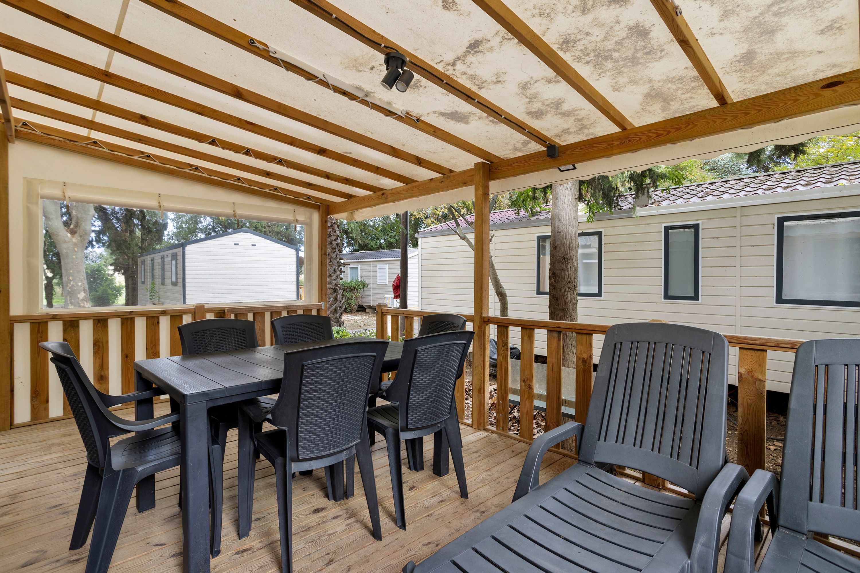 Un espacio al aire libre techado con piso de madera, sillas de plástico negro y una mesa.