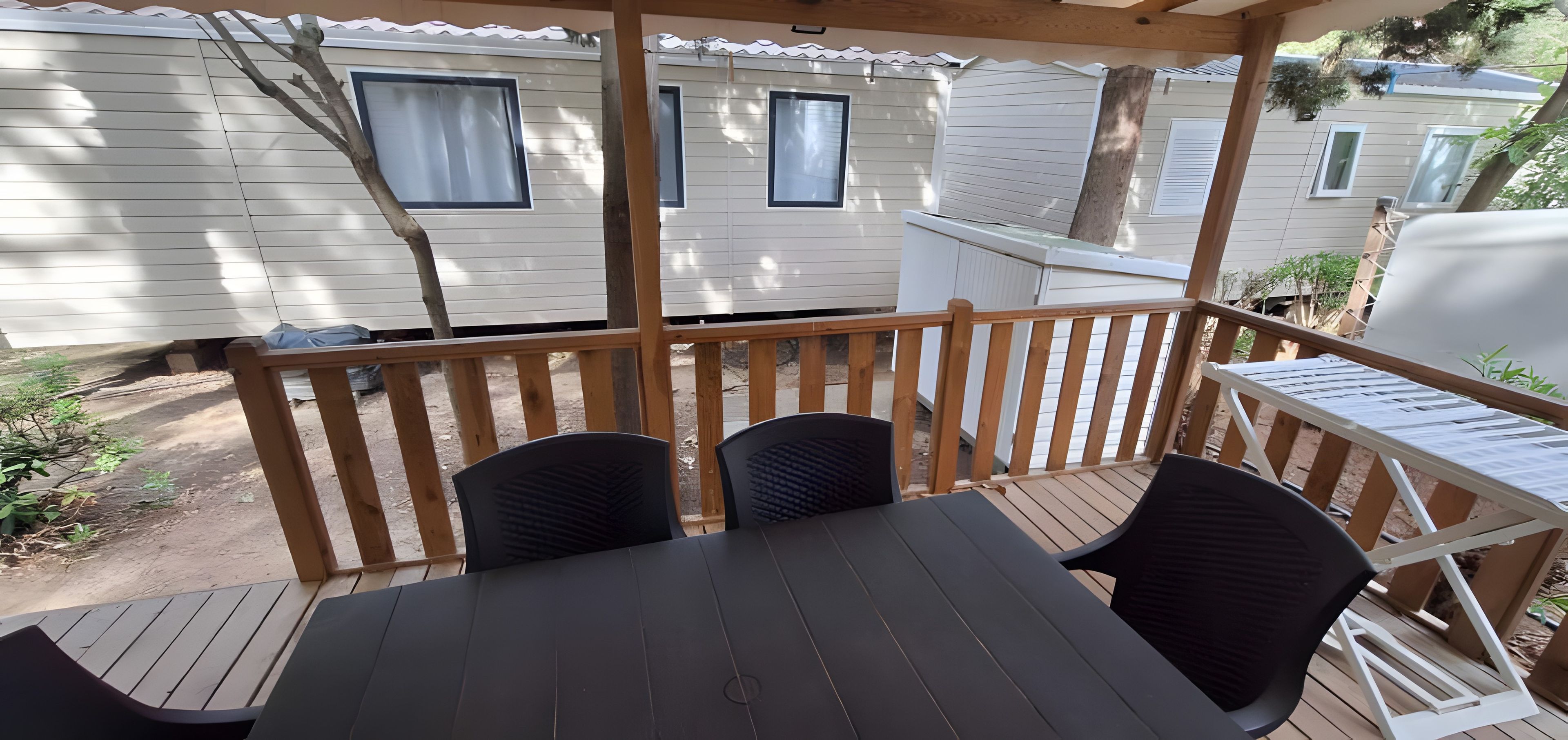 A covered outdoor seating area with a table and chairs, a wooden railing, and a view of adjacent mobile homes.