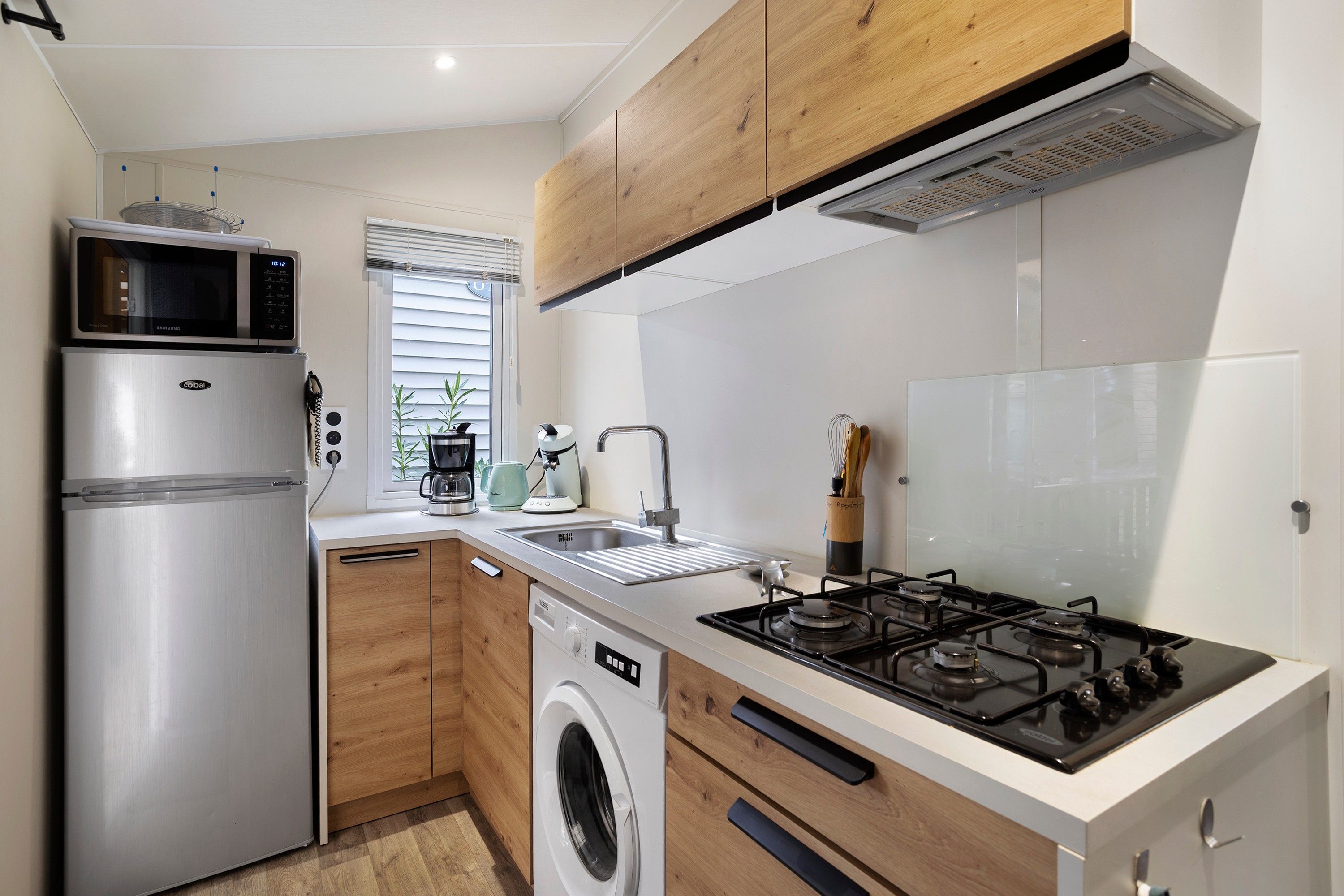 A compact kitchen with a stainless steel refrigerator, a built-in microwave, a gas stove, a washing machine, and wooden cabinets.