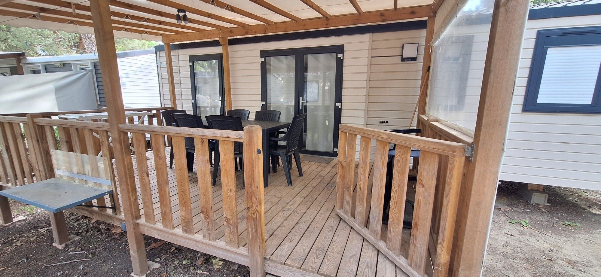Une terrasse extérieure couverte, dotée d’une table et de chaises, d’une rambarde en bois et d’une porte-fenêtre coulissante donnant sur un espace abrité.