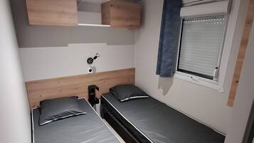 Une chambre de taille modeste, équipée de deux lits simples, d’une petite table de chevet, d’une lampe fixée au mur et d’une fenêtre avec des stores.