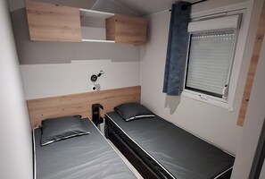 A compact room with two single beds, a small bedside table, a wall-mounted light, and a window with blinds. - Mobile home 'Bienvenue' with private terrace, Wi-Fi and air conditioning mobyl'home from 2023 (Saint-Cyprien)