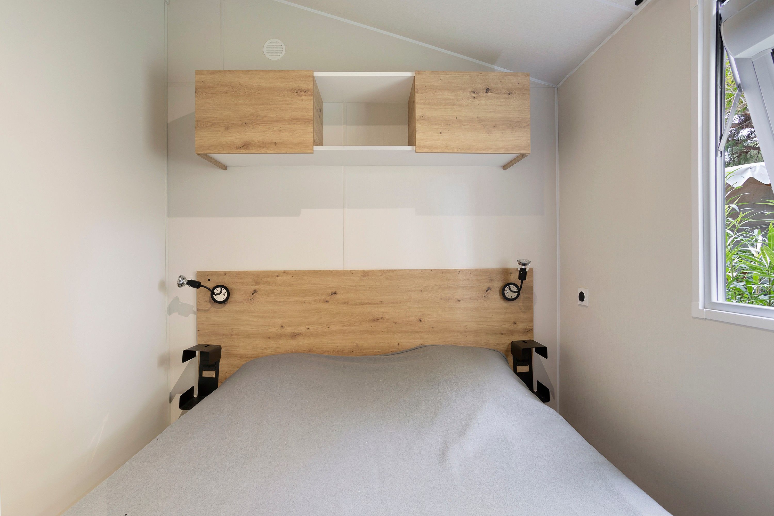 A bedroom with a wooden headboard, a bed with a grey mattress, and a window with a view of greenery.