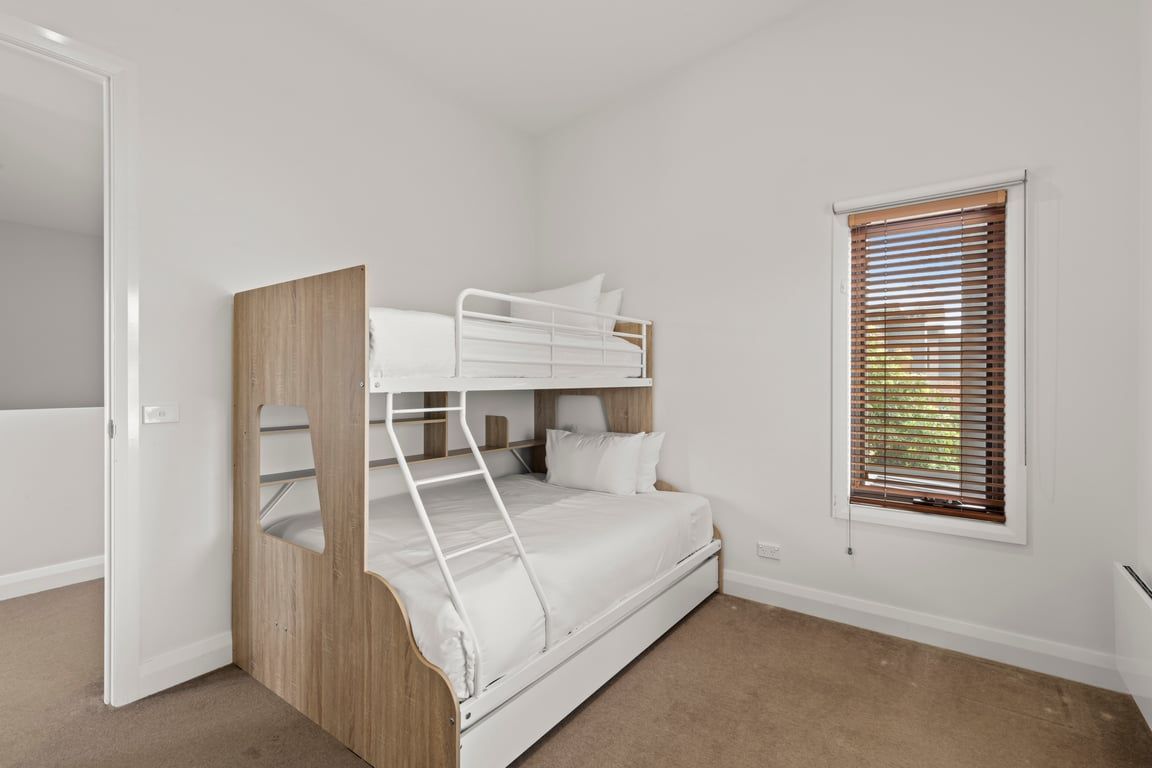 A bunk bed room with a wooden ladder, a window with blinds, and a white door.