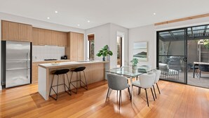 Eine moderne Küche mit Holzschränken, einem Edelstahlkühlschrank, einem Glastisch mit Barhocker und Blick durch eine Glastür auf einen Außenbereich.