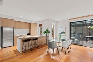 Eine moderne Küche mit Holzschränken, einem Edelstahlkühlschrank, einem Glastisch mit Barhocker und Blick durch eine Glastür auf einen Außenbereich.