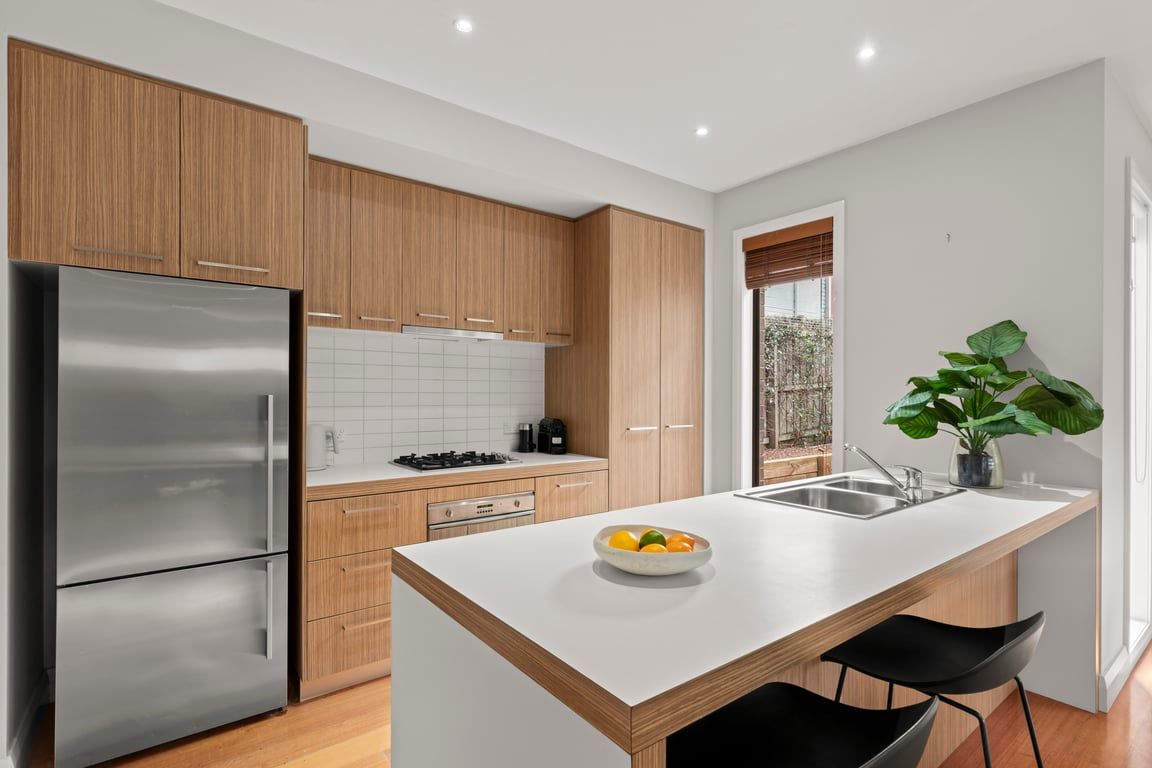 A modern kitchen with wooden cabinets, a stainless steel refrigerator, a white countertop, and a bowl of fruit.