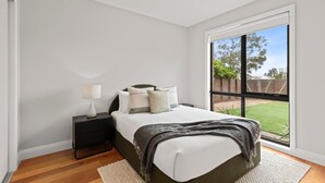 Ein Schlafzimmer mit großem Fenster, einem Bett mit Kissen, einem Nachttisch mit Lampe und Blick ins Freie.