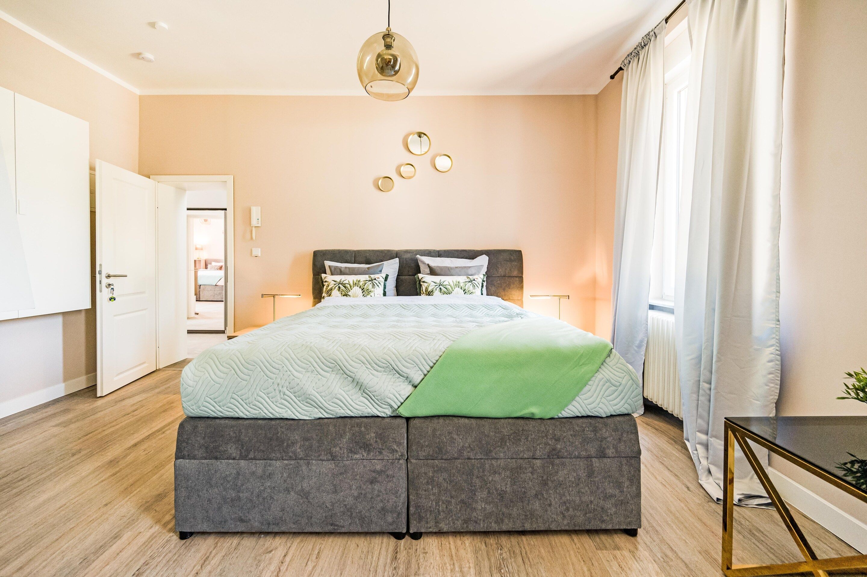 A bedroom with a bed, bedside tables, a hanging light fixture, a door, and a view of a hallway.