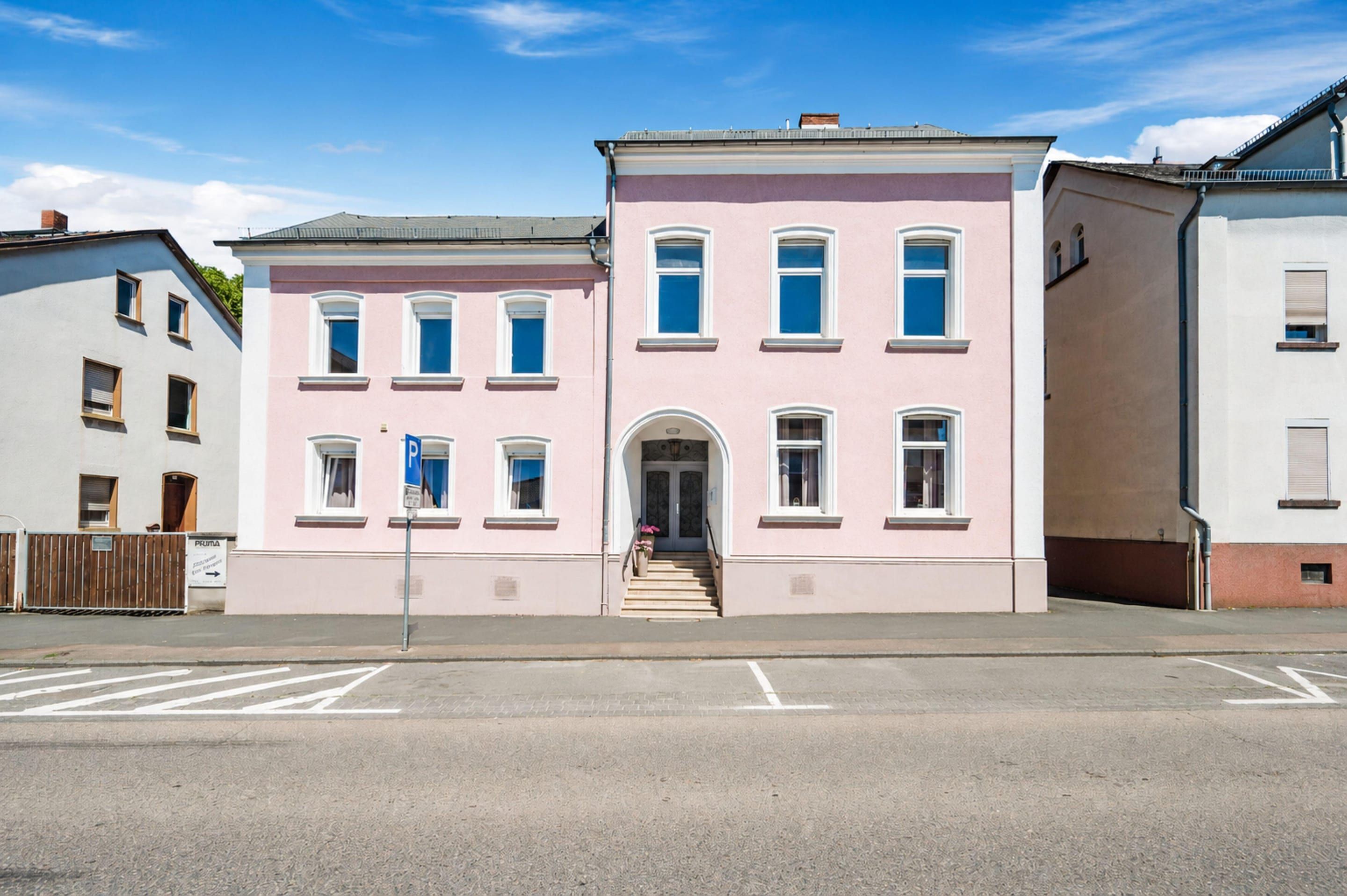 A two-story building with a pink facade, a central entrance with steps, and a parking sign.
