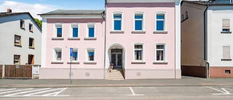 A two-story building with a pink facade, a central entrance with steps, and a parking sign.