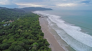 Luftaufnahme einer Küstenlandschaft mit Strand, dichtem Grün und einer fernen Bergkette.