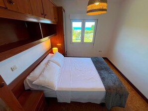 Ein Schlafzimmer mit Bett, Nachttisch, Lampe, Fenster mit Blick und einem Holzkopfteil.