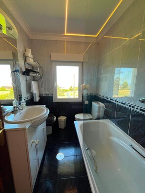 Un baño moderno con bañera, inodoro y lavamanos. Hay una ventana que proporciona luz natural y vistas a la vegetación exterior.